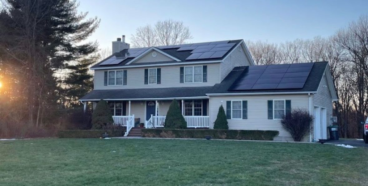 Photo-Apr-08-2025,-8-56-21-AM Two-story house with solar panels on the roof, surrounded by a lawn and trees. The sunset is visible behind the trees on the left.